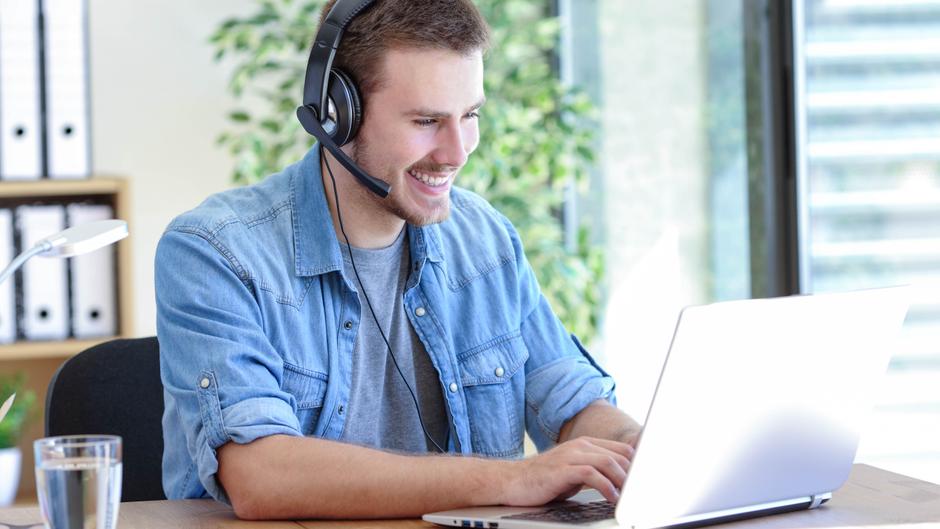 Mann mit Headset lächelt und arbeitet an einem Laptop in einem modernen Büro.