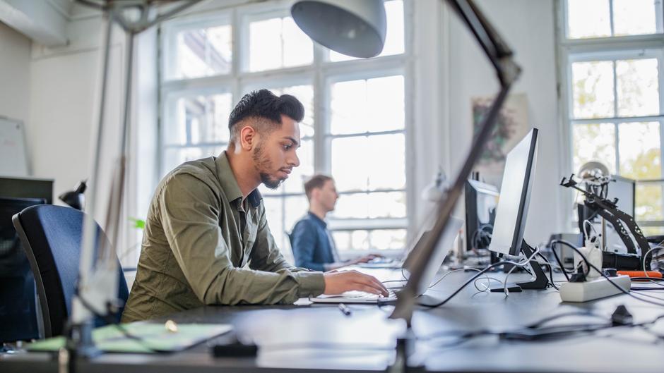 Mann arbeitet konzentriert an einem Computer in einem modernen Büro mit großen Fenstern.