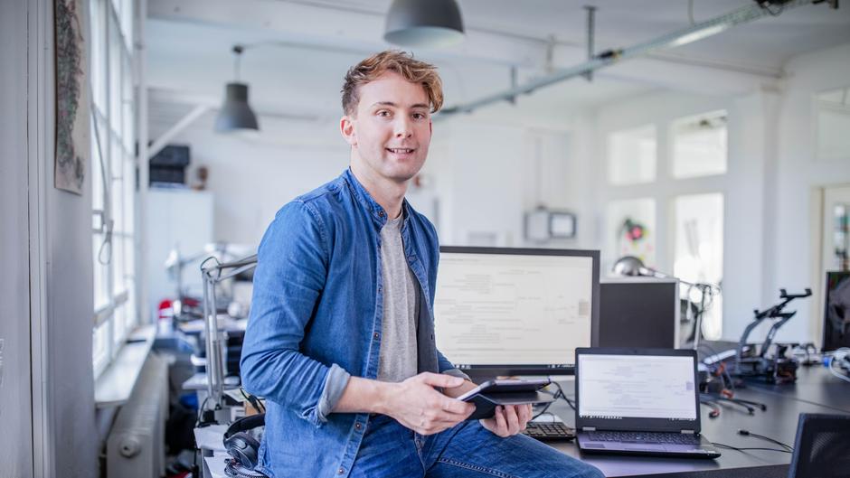 Junger Mann sitzt an einem Schreibtisch mit mehreren Computern und hält ein Tablet in der Hand.
