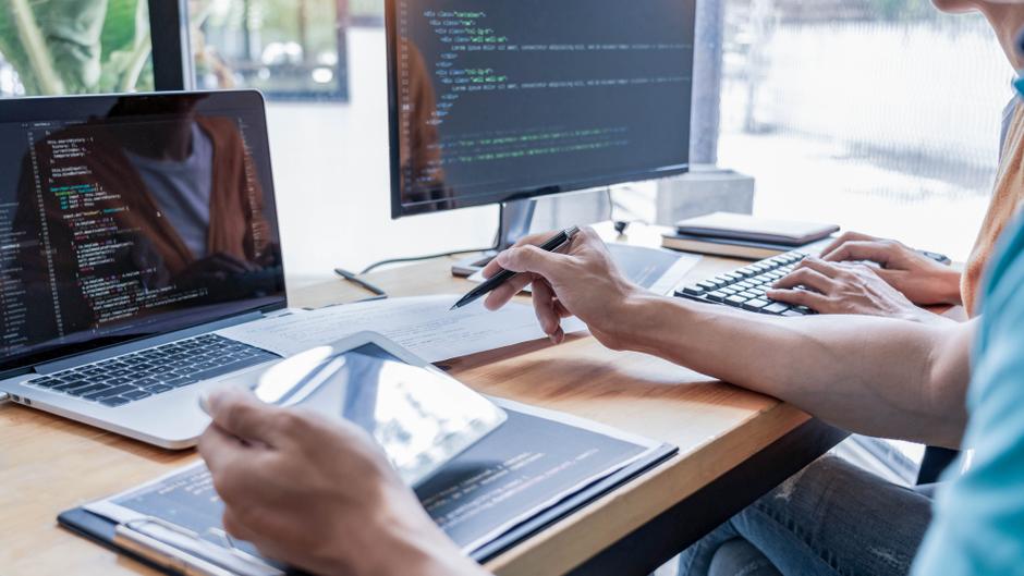 Zwei Personen arbeiten an Laptops und einem Monitor, während sie Programmiercode und Notizen verwenden.