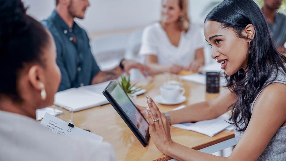 Eine Frau zeigt einem Mann auf einem Tablet während eines Meetings, im Hintergrund sind weitere Personen und Unterlagen sichtbar.