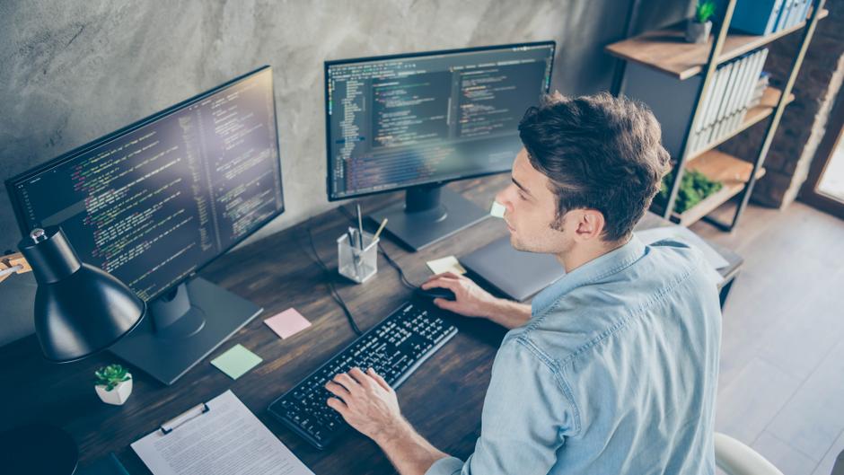Ein Mann sitzt an einem Schreibtisch mit zwei Monitoren, auf denen Programmiercode angezeigt wird.