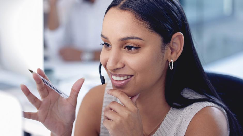 Frau mit Headset lächelt und hält einen Stift in der Hand, während sie am Computer arbeitet.