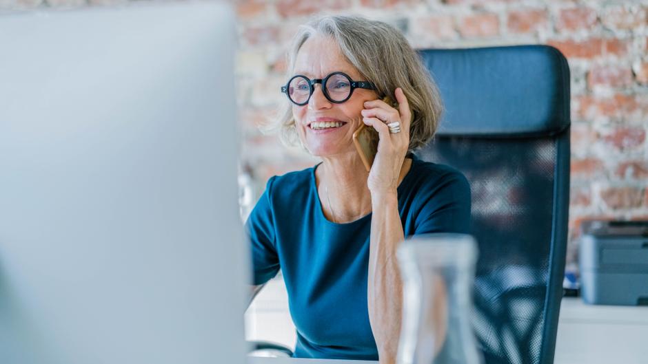 Ältere Frau mit Brille spricht am Telefon, sitzt an einem Schreibtisch mit Computer und Wasserkrug.