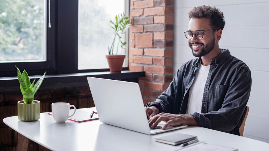 Mann mit Brille sitzt an einem Tisch und arbeitet an einem Laptop, neben ihm eine Tasse und eine Pflanze.