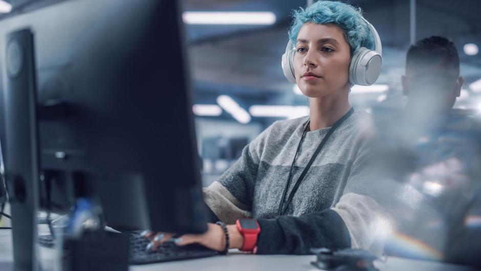 Junge Person mit blauen Haaren und Kopfhörern arbeitet am Computer in einem modernen Büro.