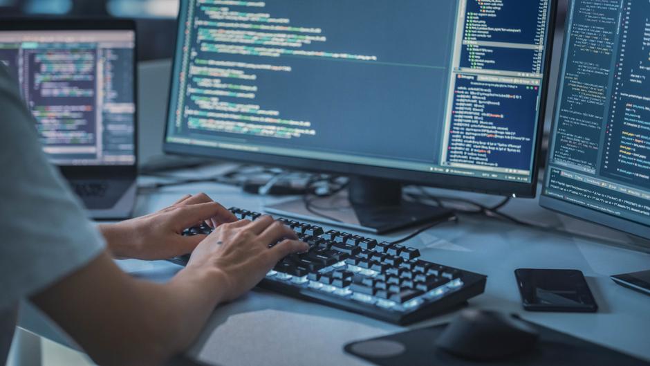 Hände tippen auf einer beleuchteten Tastatur, während zwei Monitore mit Programmiercode angezeigt werden.