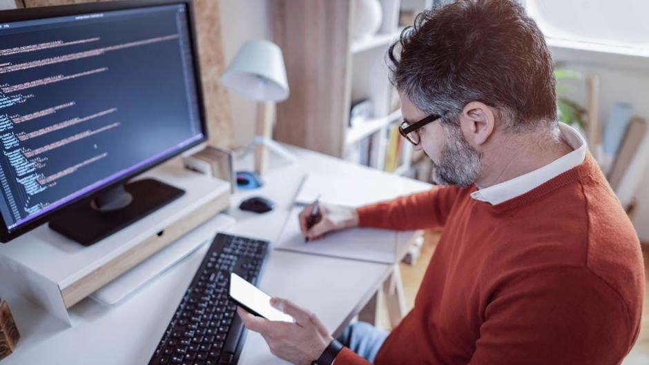 Mann in einem roten Pullover sitzt am Schreibtisch, schreibt auf Papier und hält ein Smartphone in der Hand.