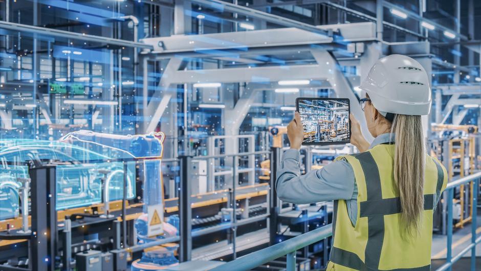 Ingenieurin mit Schutzhelm und Warnweste hält ein Tablet in einer modernen Fabrik mit holografischen Darstellungen.