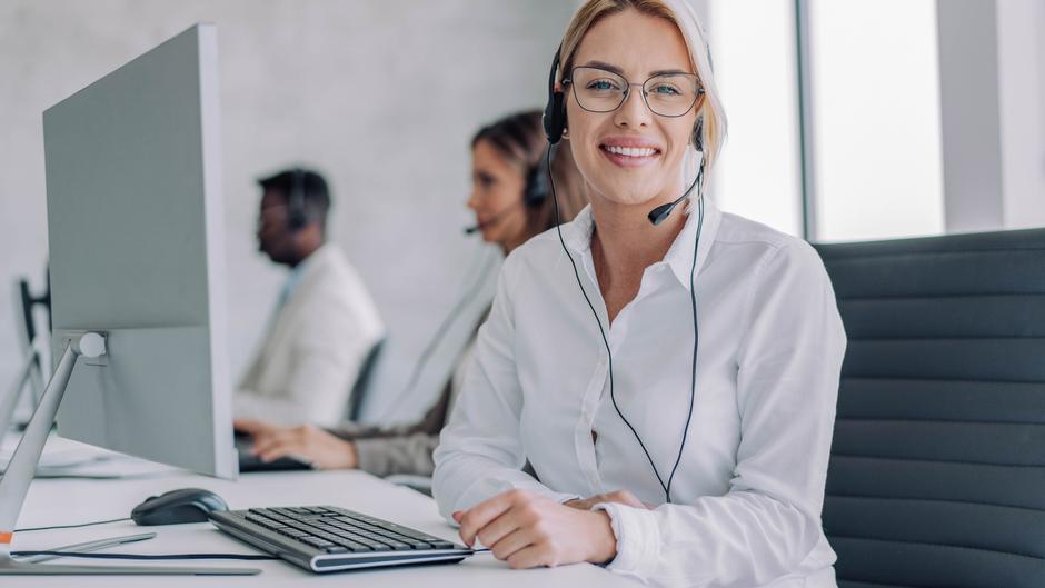 Lächelnde Frau mit Headset sitzt an einem Schreibtisch mit Computer und Tastatur, im Hintergrund weitere Personen.