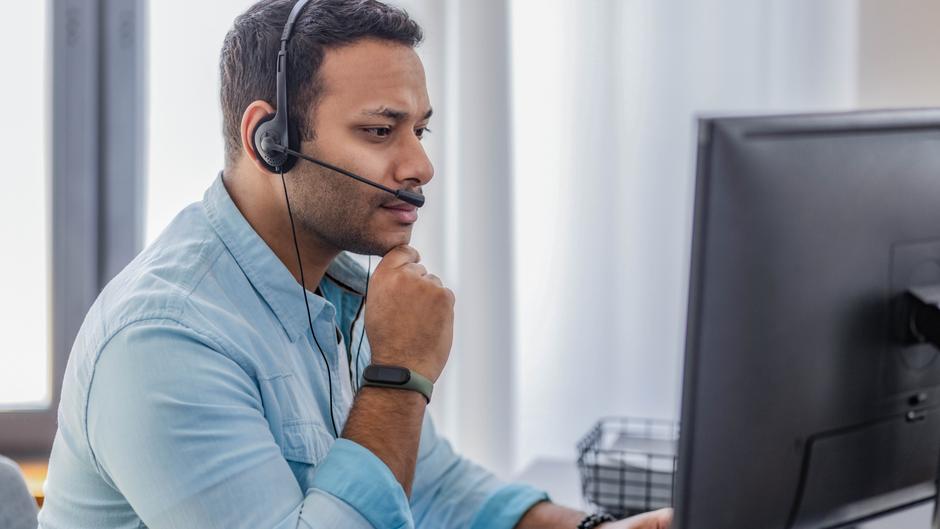Mann mit Headset sitzt vor einem Computer und schaut nachdenklich auf den Bildschirm.