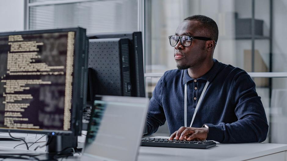 Ein Mann mit Brille sitzt an einem Schreibtisch und arbeitet konzentriert an einem Computer mit Programmiercode.