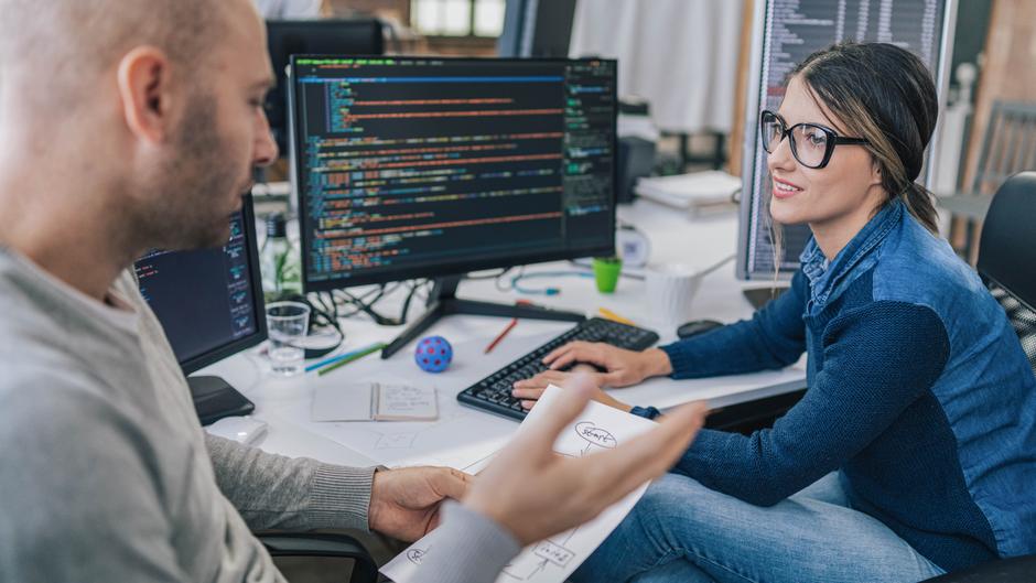 Zwei Personen sitzen an einem Schreibtisch mit Computern, diskutieren und betrachten ein Blatt Papier mit Diagrammen.