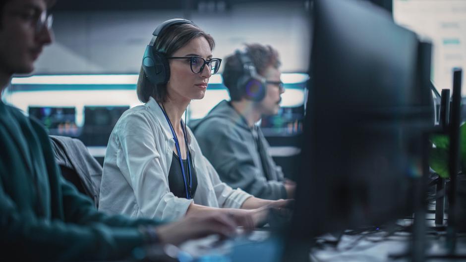 Drei Personen sitzen an Computern in einem modernen Arbeitsumfeld, eine trägt Kopfhörer und schaut konzentriert auf den Bildschirm.
