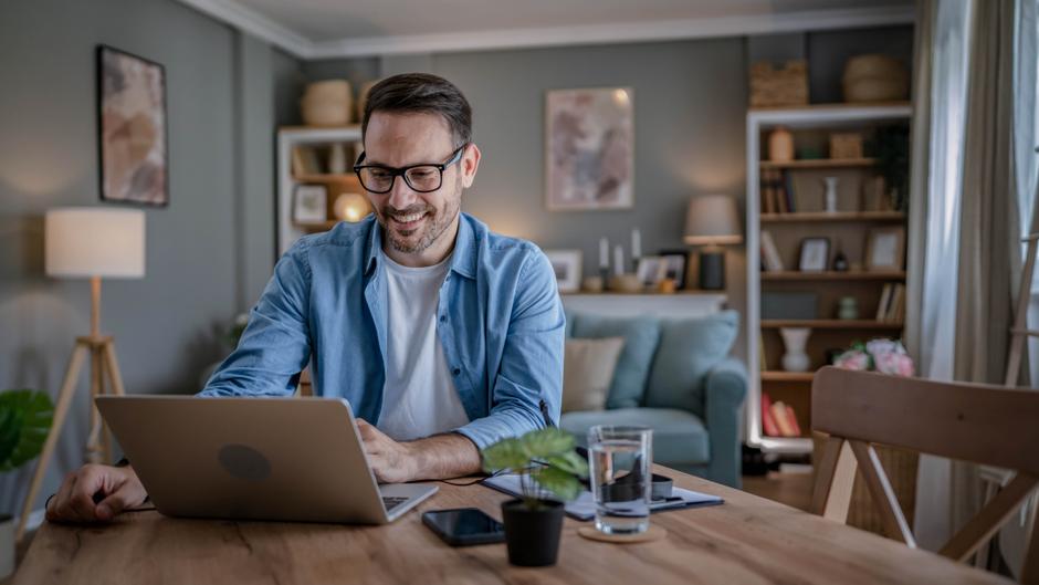 Mann mit Brille sitzt an einem Tisch und arbeitet am Laptop, im Hintergrund ein gemütliches Wohnzimmer.