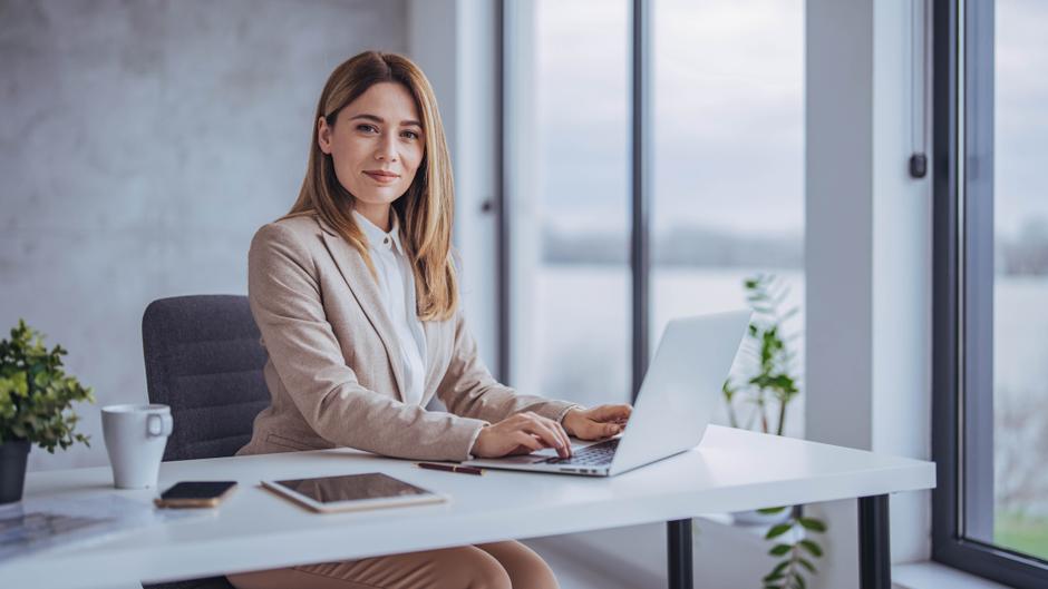 Frau im Business-Outfit sitzt an einem Schreibtisch mit Laptop, Tasse und Pflanzen im Hintergrund.