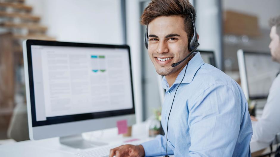 Lächelnder Mann mit Headset sitzt an einem Schreibtisch vor einem Computerbildschirm.