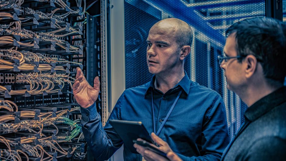 Zwei Männer stehen vor einem Serverrack mit Kabeln und diskutieren, während einer ein Tablet in der Hand hält.