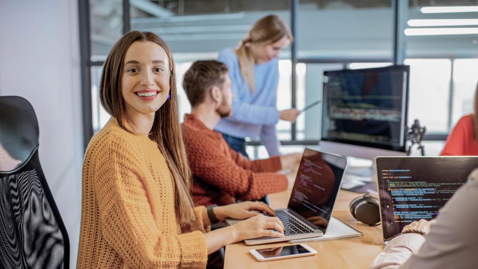 Lächelnde Frau in einem modernen Büro, die an einem Laptop arbeitet, während Kollegen im Hintergrund diskutieren.