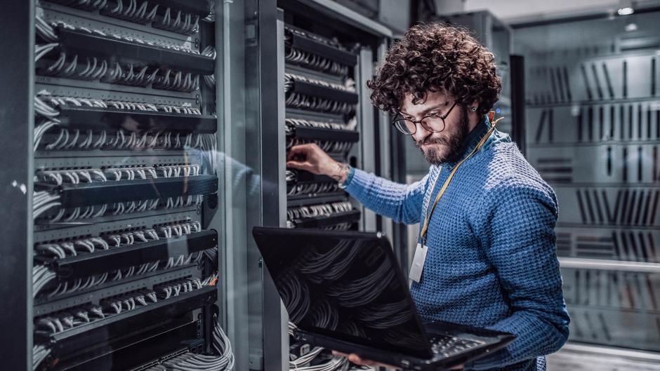 Mann mit lockigem Haar arbeitet an einem Serverrack und hält einen Laptop in der Hand.
