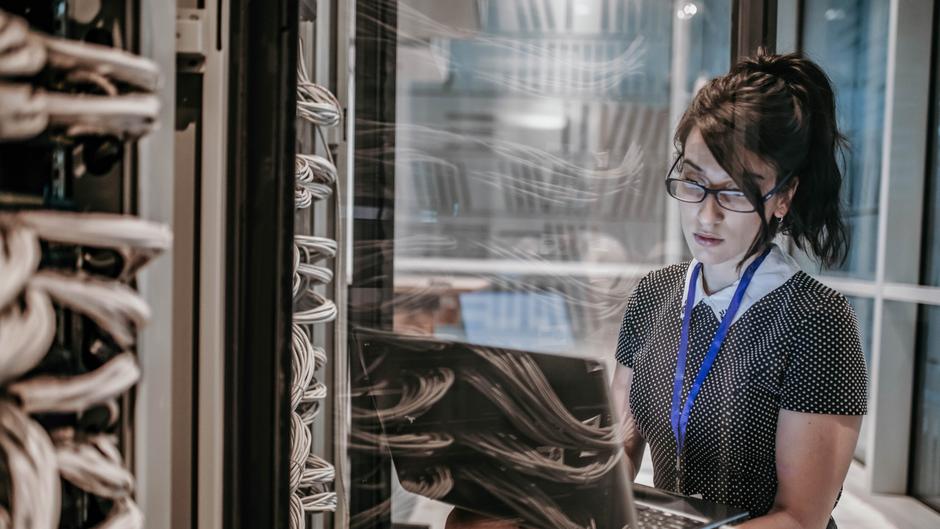 Frau mit Brille arbeitet an einem Laptop in einem Serverraum mit Kabeln im Hintergrund.