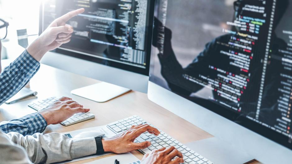 Zwei Personen arbeiten an Computern, während eine Person auf den Bildschirm zeigt, der Programmiercode anzeigt.