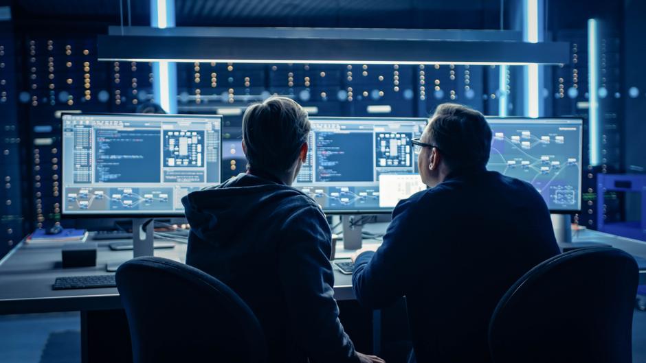 Zwei Personen sitzen an einem Tisch vor mehreren Monitoren mit Programmiercode und Diagrammen in einem dunklen Raum.