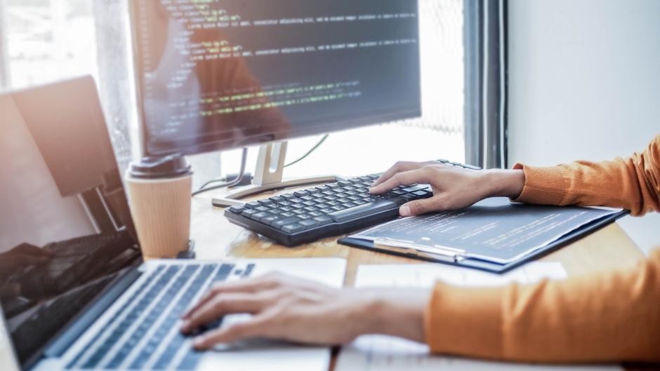 Hände tippen auf einer Tastatur, während ein Laptop und ein Monitor mit Code sichtbar sind. Ein Kaffeebecher steht auf dem Tisch.