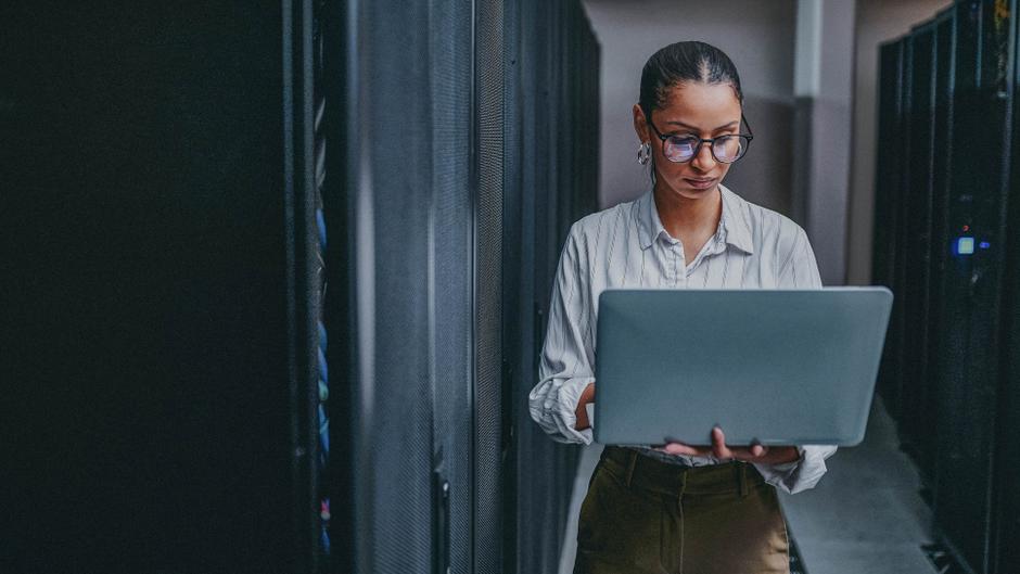 Eine Person mit Brille steht in einem Serverraum und arbeitet an einem Laptop.