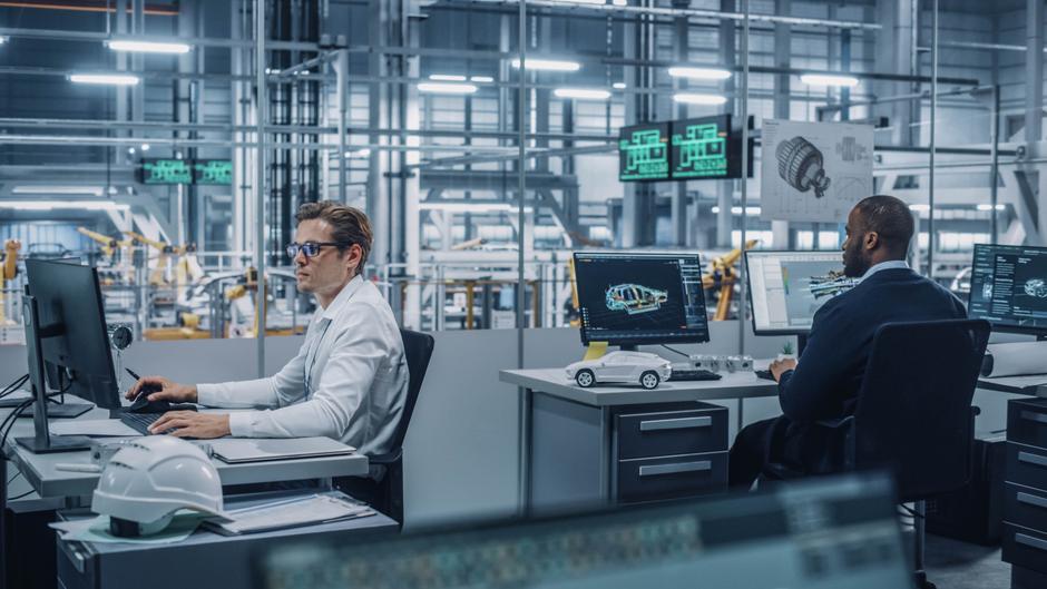 Zwei Ingenieure arbeiten an Computern in einem modernen Büro mit technischen Zeichnungen und einem Modell eines Autos.