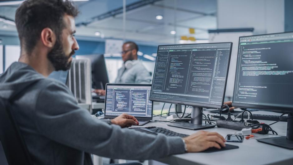 Mann arbeitet an mehreren Bildschirmen mit Programmiercode in einem modernen Büro.