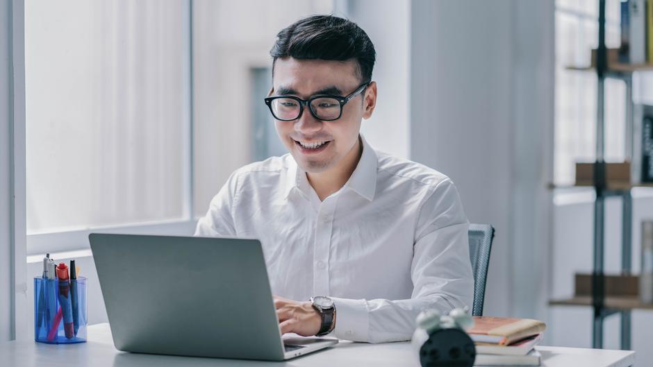 Mann mit Brille lächelt, während er an einem Laptop arbeitet, umgeben von Schreibutensilien und Büchern.