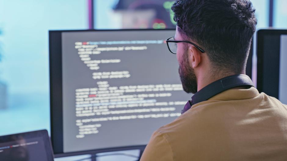 Mann mit Brille und Kopfhörern schaut auf einen Computerbildschirm mit Codezeilen.