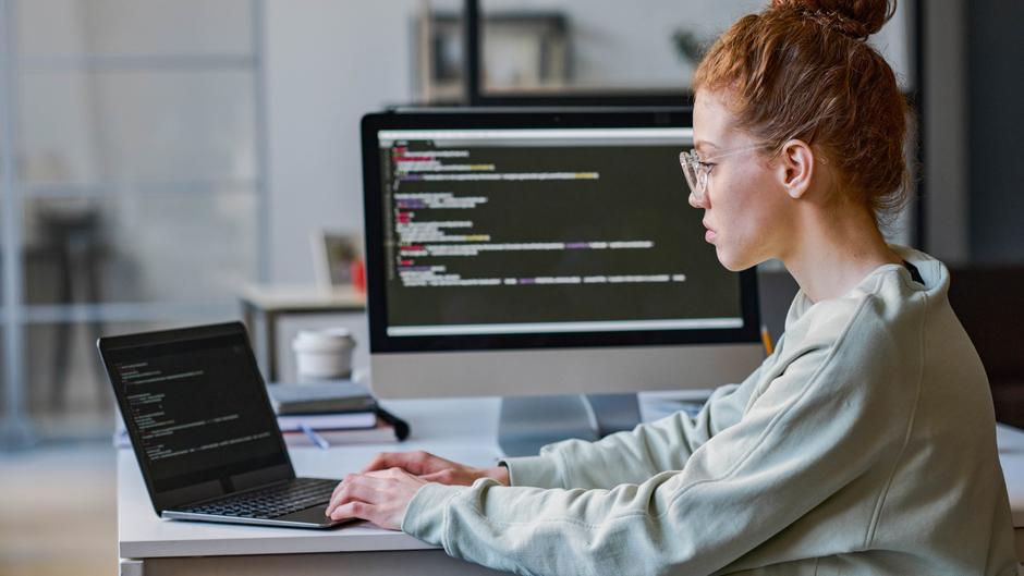 Eine Person mit Brille arbeitet konzentriert an einem Laptop, während ein Monitor mit Code sichtbar ist.
