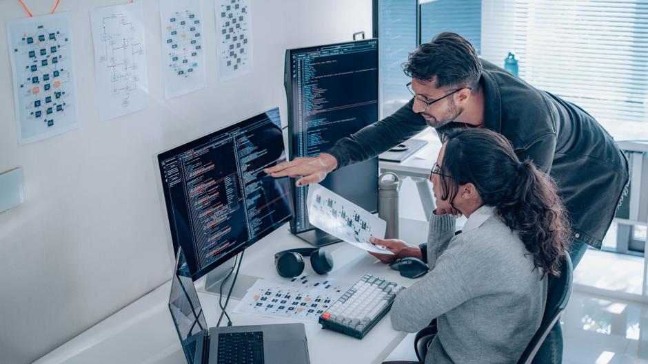 Zwei Personen arbeiten an einem Schreibtisch mit mehreren Monitoren, auf denen Code und Diagramme angezeigt werden.
