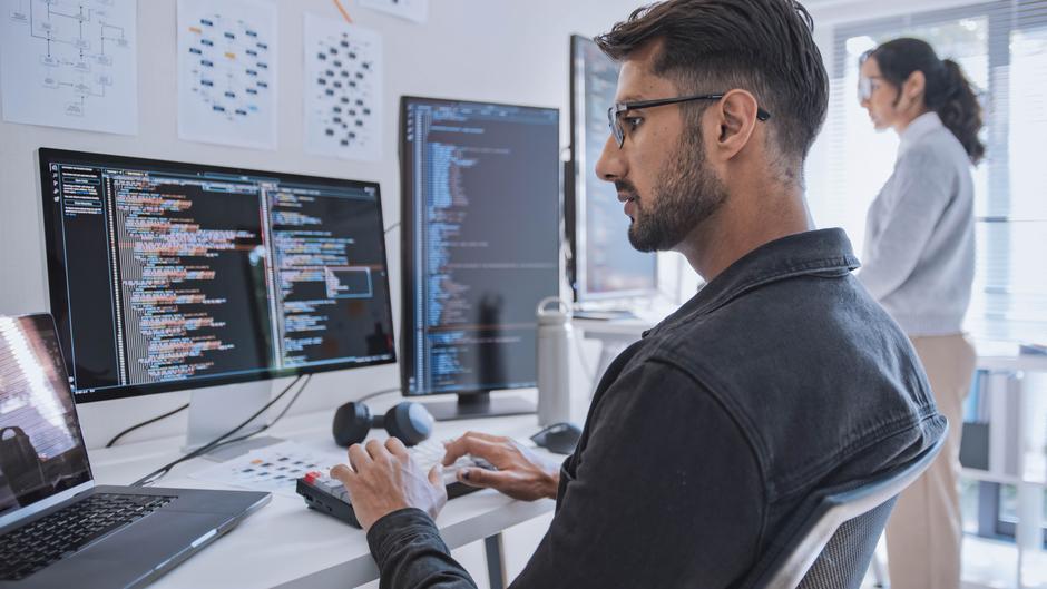 Ein Mann sitzt an einem Schreibtisch und arbeitet an mehreren Monitoren mit Programmiercode, während eine Frau im Hintergrund steht.