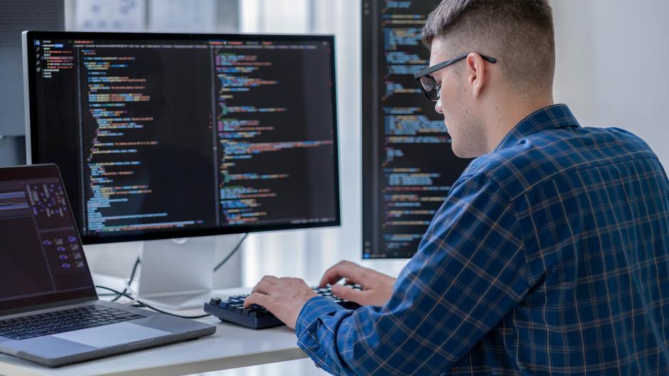 Ein Mann in einem karierten Hemd tippt an einer Tastatur vor zwei Monitoren mit Programmiercode.