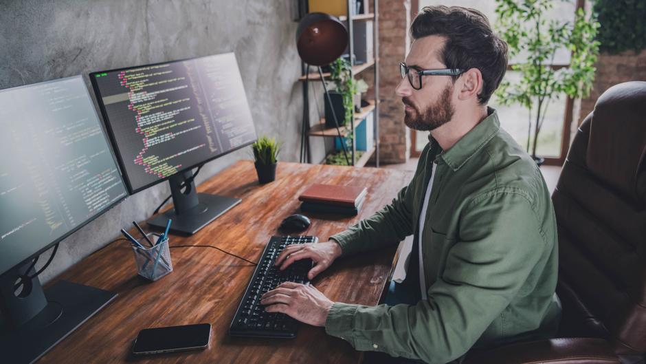 Mann mit Brille sitzt an einem Schreibtisch und tippt auf einer Tastatur, zwei Monitore zeigen Programmiercode.
