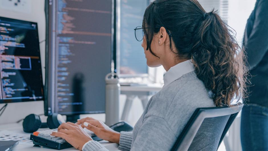 Eine Person mit Brille tippt an einer Tastatur, während sie auf zwei Computerbildschirme mit Code schaut.