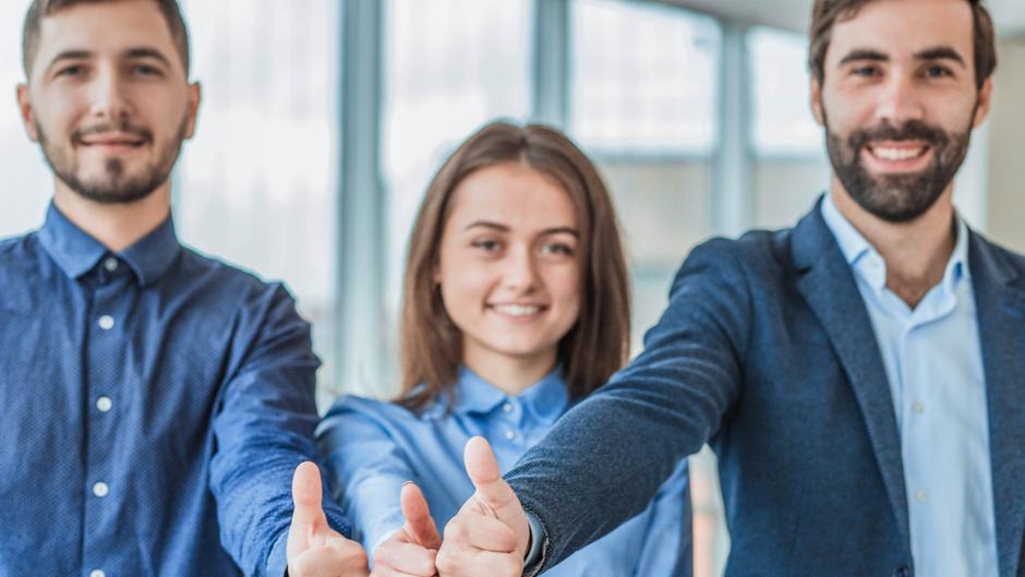 Drei Personen zeigen lächelnd Daumen hoch in einem modernen Büro mit großen Fenstern.