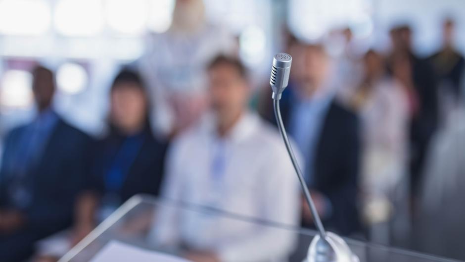 Mikrofon im Vordergrund mit unscharfem Publikum im Hintergrund während einer Präsentation.
