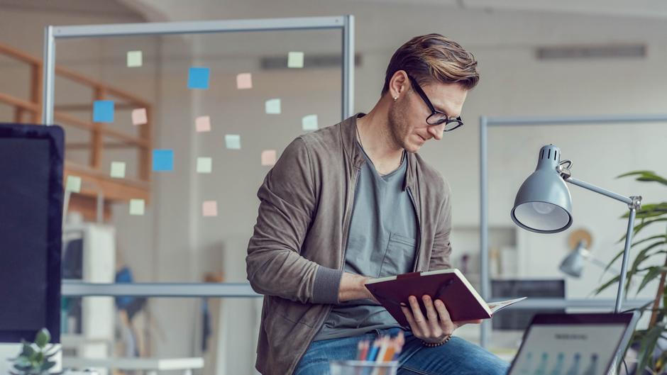 Mann mit Brille sitzt in einem modernen Büro und liest in einem Notizbuch, im Hintergrund bunte Haftnotizen.
