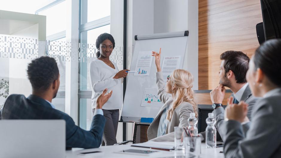Eine Person präsentiert in einem Meetingraum Grafiken auf einem Flipchart, während andere Teilnehmer aufmerksam zuhören.