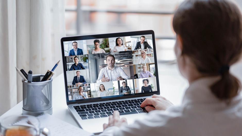 Eine Person sitzt vor einem Laptop, auf dem eine Videokonferenz mit mehreren Teilnehmern zu sehen ist.