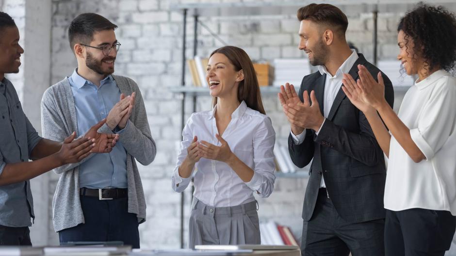 Sechs Personen in einem modernen Büro applaudieren und lachen, während eine Frau in der Mitte spricht.