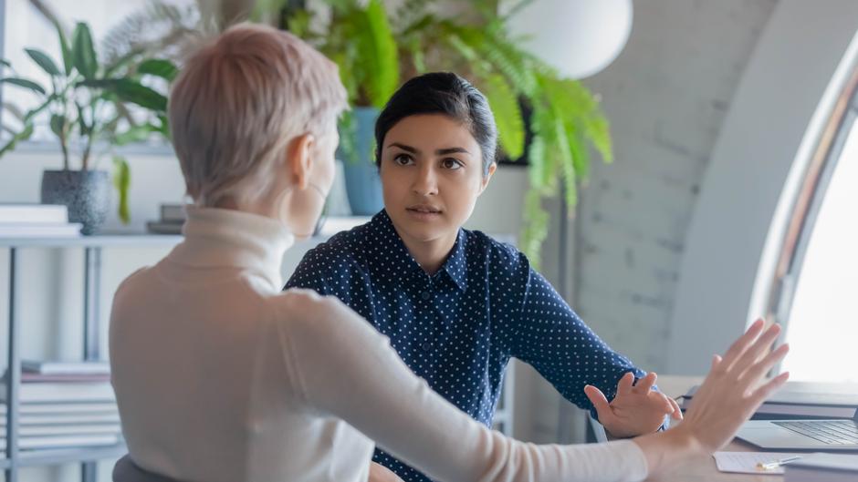 Zwei Frauen sitzen an einem Tisch und führen ein intensives Gespräch, eine gestikuliert. Pflanzen sind im Hintergrund sichtbar.