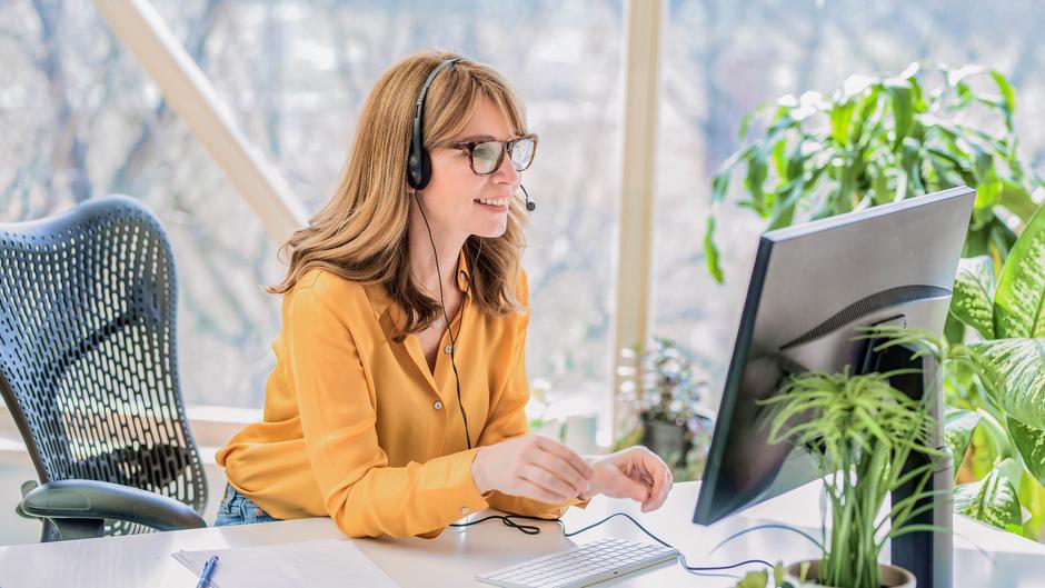 Frau mit Headset sitzt an einem Schreibtisch und lächelt, während sie auf einen Computerbildschirm schaut.