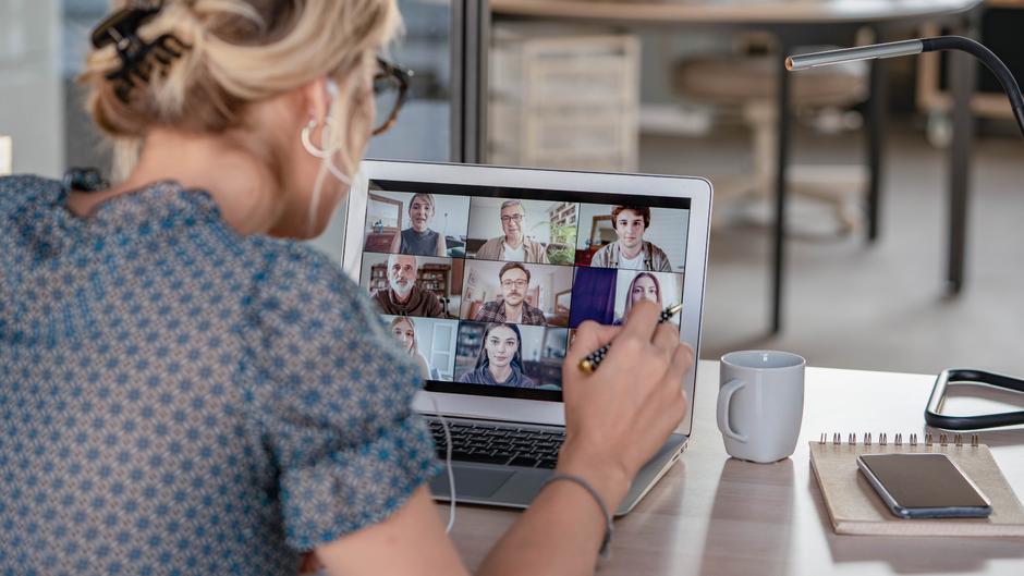 Eine Person mit Kopfhörern sitzt vor einem Laptop, auf dem ein Videoanruf mit mehreren Teilnehmern zu sehen ist.
