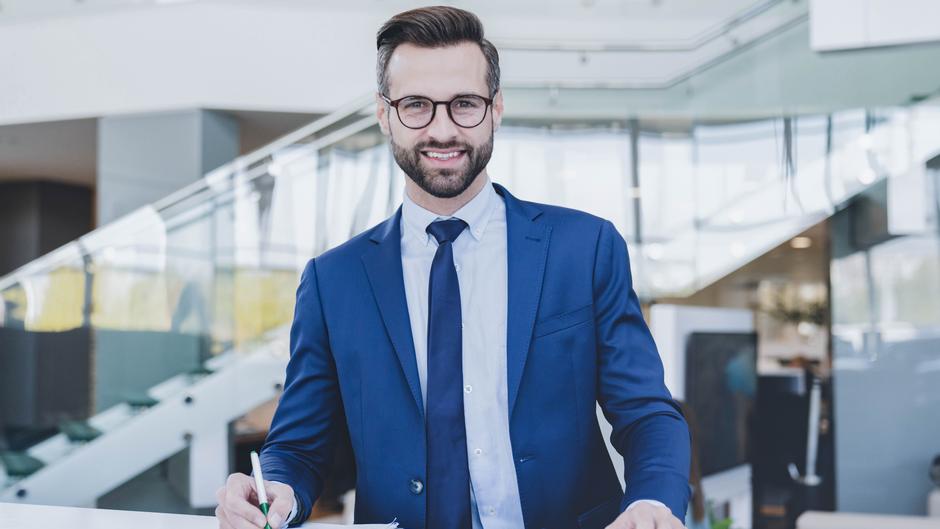 Mann in Anzug mit Brille lächelt am Schreibtisch und hält einen Stift in der Hand.