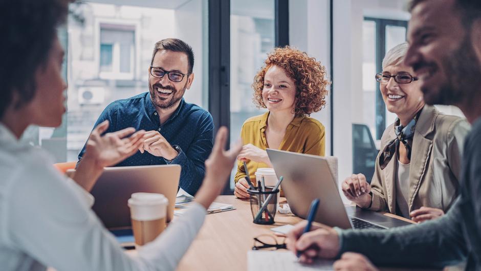 Fünf Personen in einem Besprechungsraum lachen und diskutieren, Laptops und Kaffeebecher auf dem Tisch.